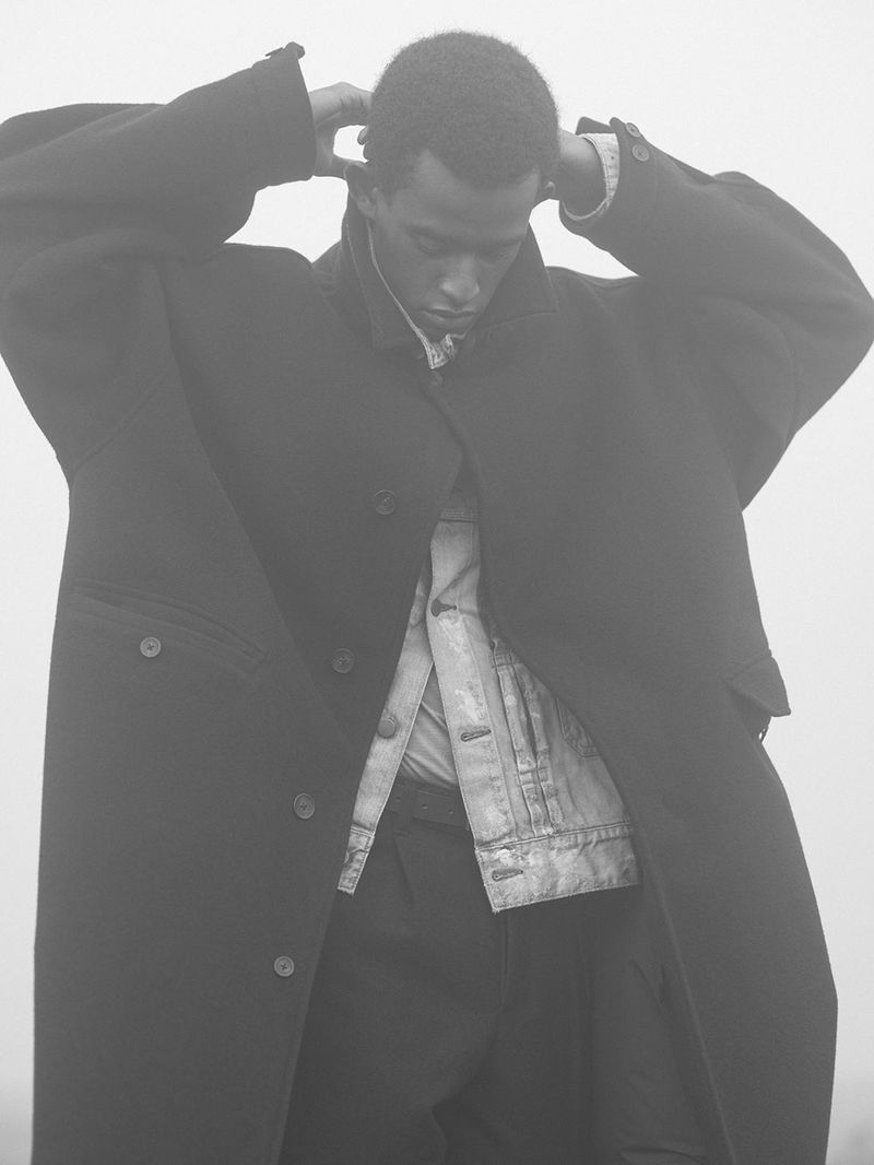 Portrait of an adult Black male in black overcoat adjusting collar with an unbuttoned jean jacket in a muted, foggy outdoor setting.