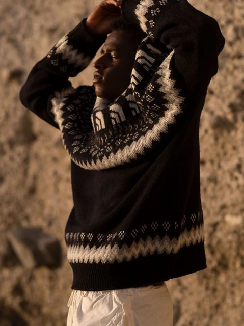 An adult male poses with his hands behind his head in front of a textured rock wall, wearing a patterned knit sweater and light trousers.
