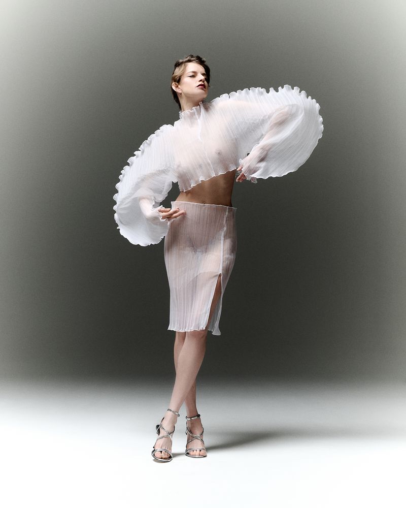 A slender woman strikes a pose in a translucent white, ruffled top and skirt set against a muted grey background in a studio setting.