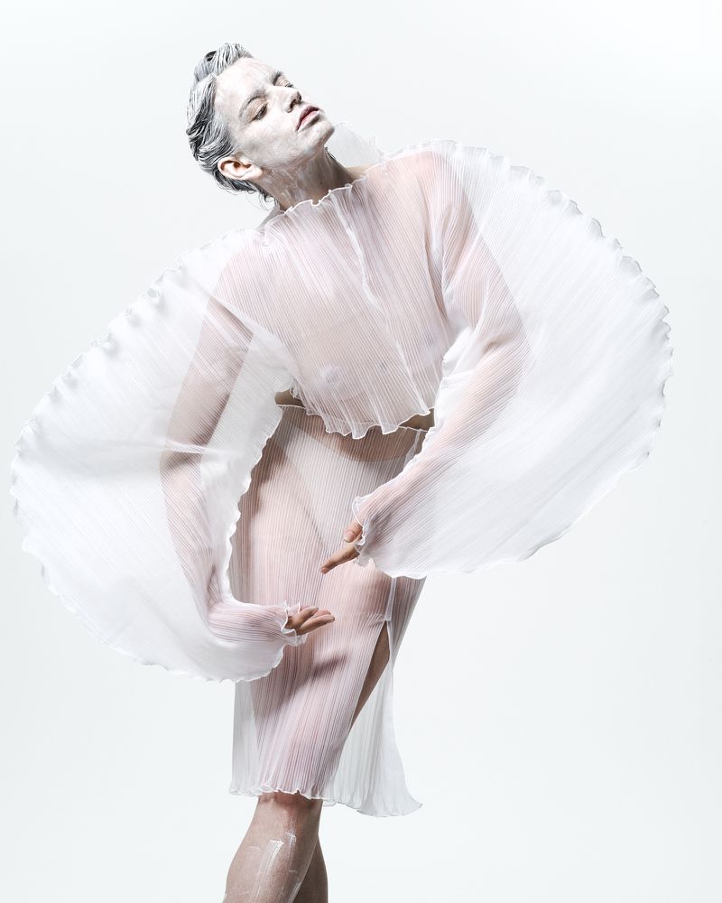Artistic fashion shot of a woman covered in white paint, wearing a white dress, and posing in a bright studio with a light background.