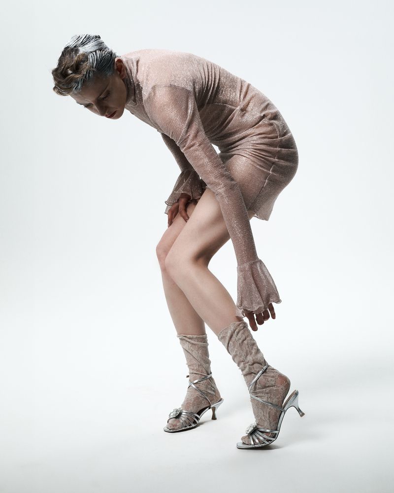 Woman in sparkling dress bending over in a studio, wearing socks and heels. Silver hair, neutral makeup, studio lighting, fashion shot.