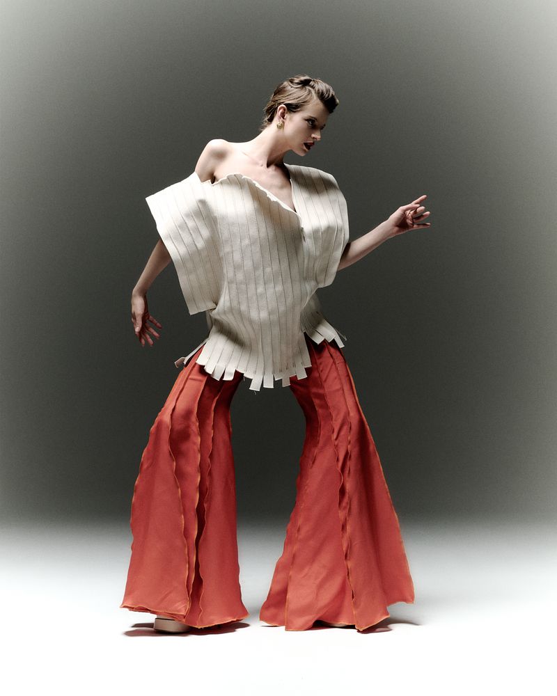 Fashion photo of a woman wearing a uniquely designed white top and orange pants in a studio setting with neutral background.