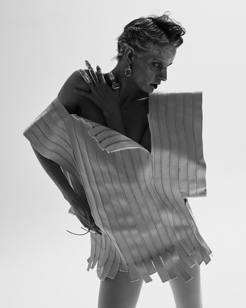 Black and white fashion portrait of a person with short hair wearing a structured garment with large earrings and multiple rings.
