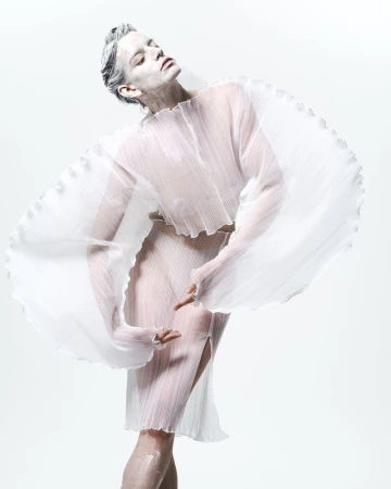 Model in white pleated sheer garment posing against a white background with white-painted skin and hair