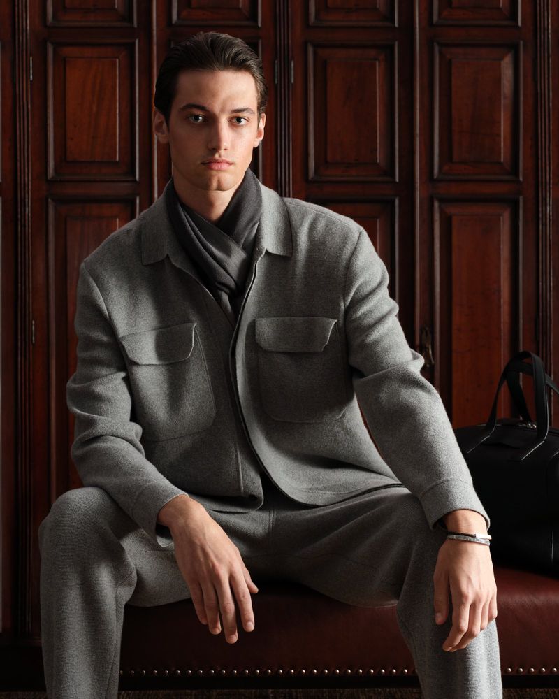 Male model poses in gray knitwear jacket and pants with a dark scarf indoors, sitting on a brown leather bench against wood paneling.
