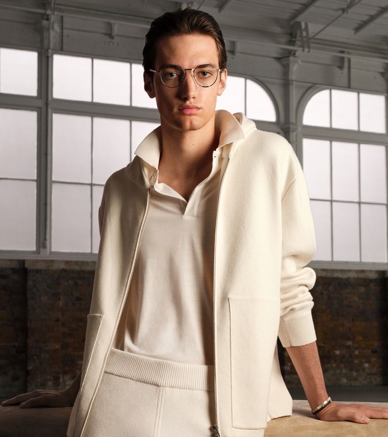 Man posing indoors in an off-white zip-up jacket, polo shirt and pants. He is wearing glasses and leans on a neutral-toned surface.