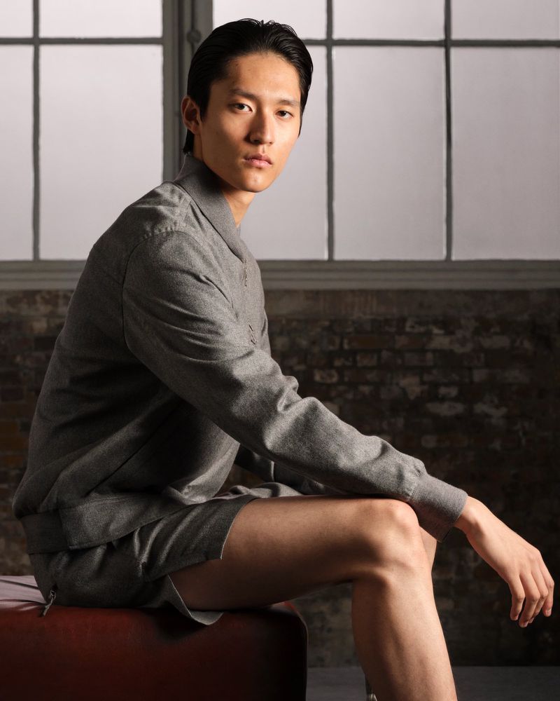Young Asian male model in grey matching set, sitting indoors near window. Portrait with neutral expression, medium shot, studio lighting.