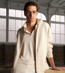 A young male model wearing cream knitwear and wire-rimmed glasses in an interior space with large arched windows.
