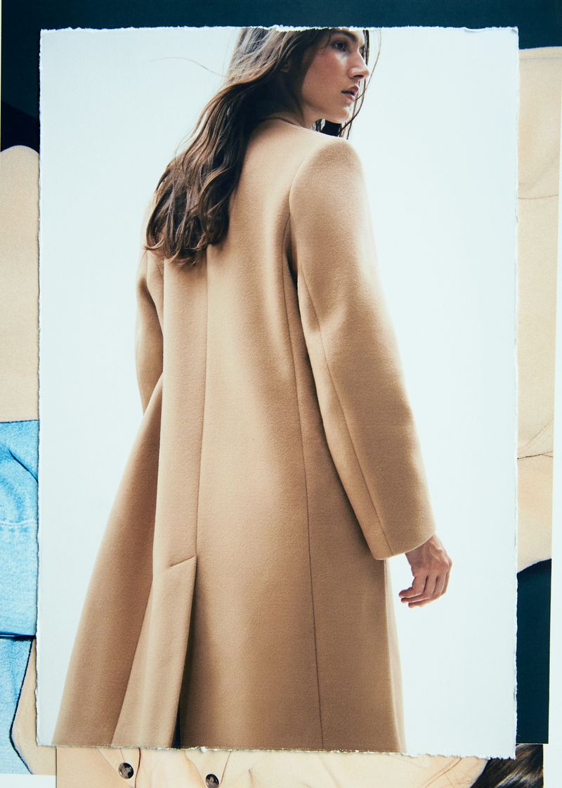 A woman with long brown hair poses wearing a tan coat on a white backdrop with framed layers of textile-like materials.