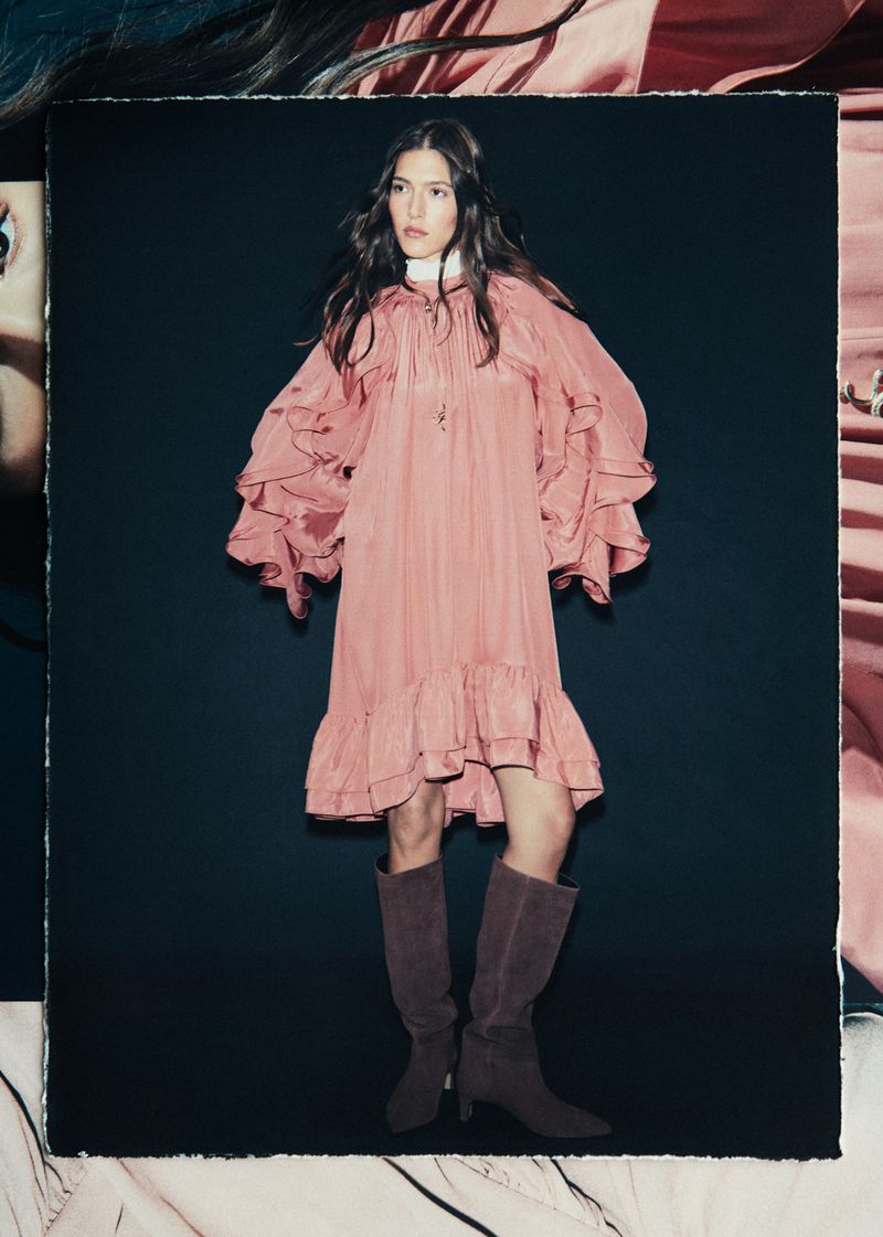 A woman poses wearing a ruffled pink dress and knee-high boots against a dark backdrop in a studio fashion photograph.