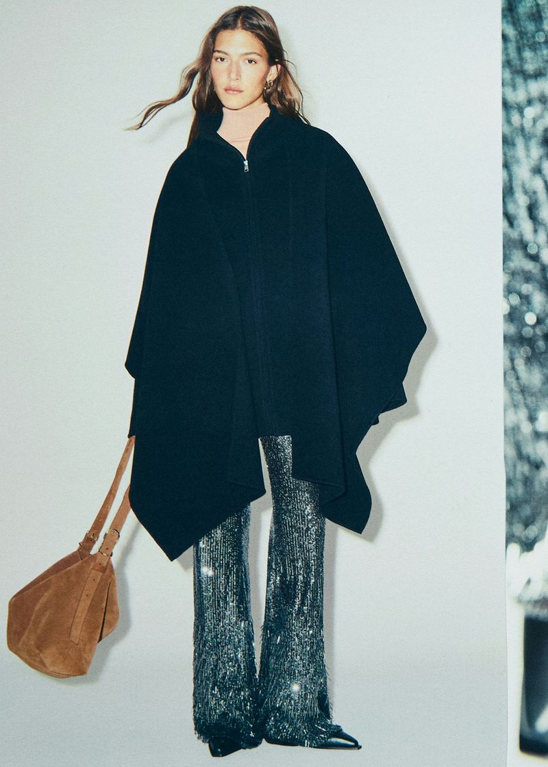 Full-length portrait of a woman wearing a black cape, sequin pants, and carrying a brown bag, posed in a bright, neutral studio.