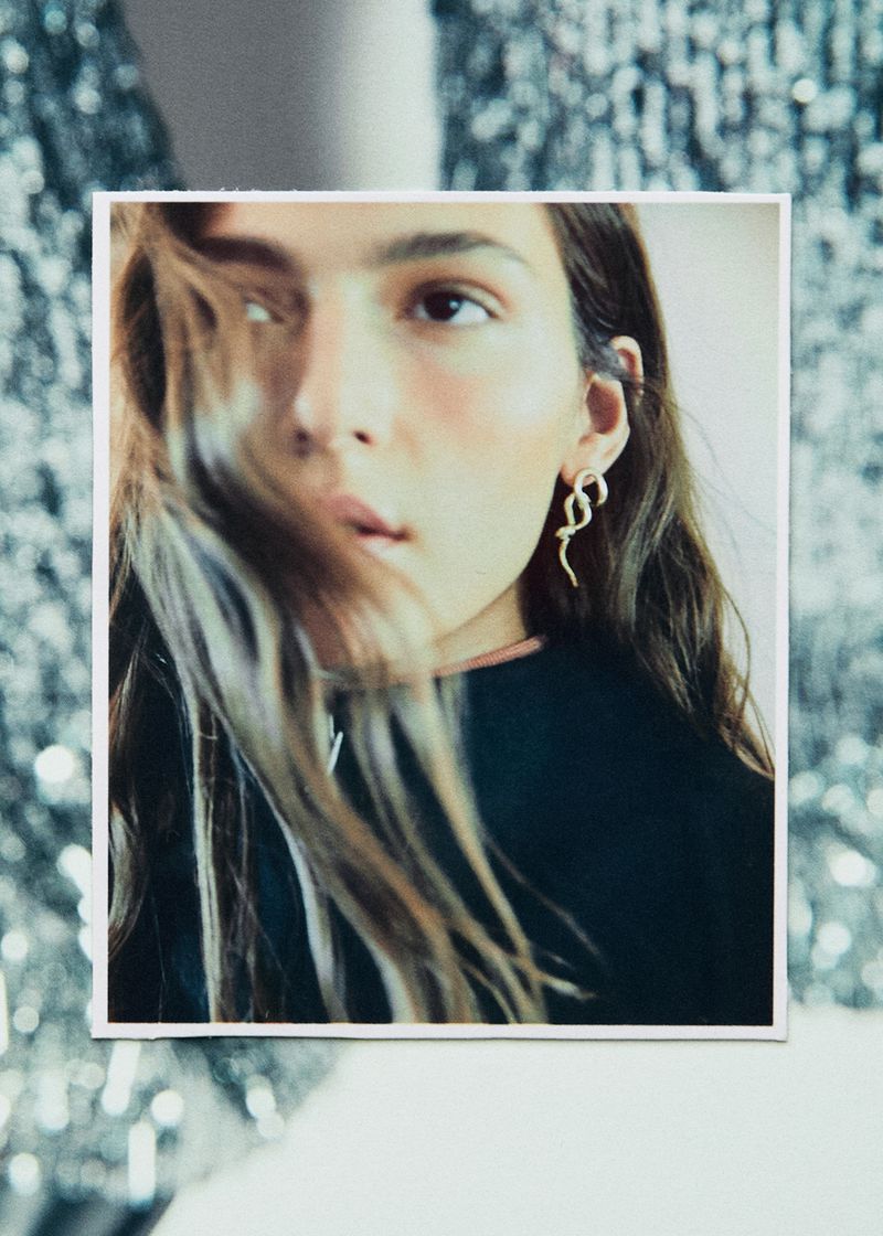 Close-up of a young adult woman in a black top wearing gold snake earrings, with a blurred shimmering background.