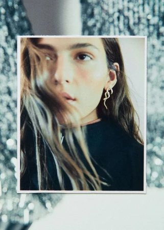 Close-up of a young adult woman in a black top wearing gold snake earrings, with a blurred shimmering background.