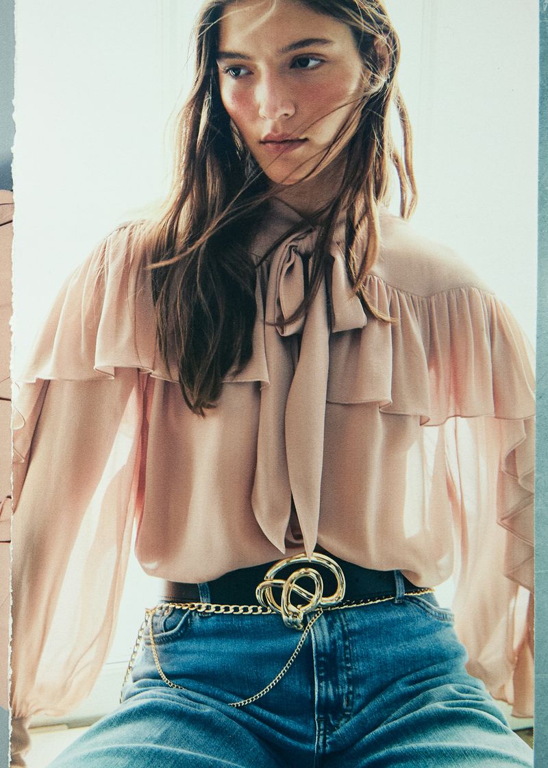 Fashion shot of a female model wearing a light pink blouse with ruffles, jeans, and a decorative gold belt. Indoors with soft lighting.