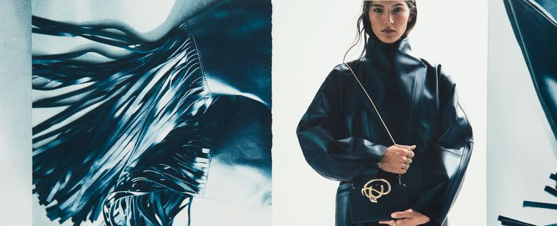 Fashion editorial shot of a woman in black leather attire with a gold chain purse, next to a jacket with fringe, set against a white backdrop.