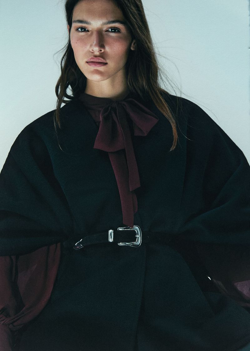 Close-up of a female model wearing a black poncho belted at the waist with a large silver buckle, and a chiffon bow-tie blouse.