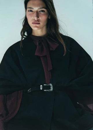 Close-up of a female model wearing a black poncho belted at the waist with a large silver buckle, and a chiffon bow-tie blouse.