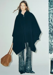 Model in black cape and sparkly trousers holding a brown suede bag against a white studio background.