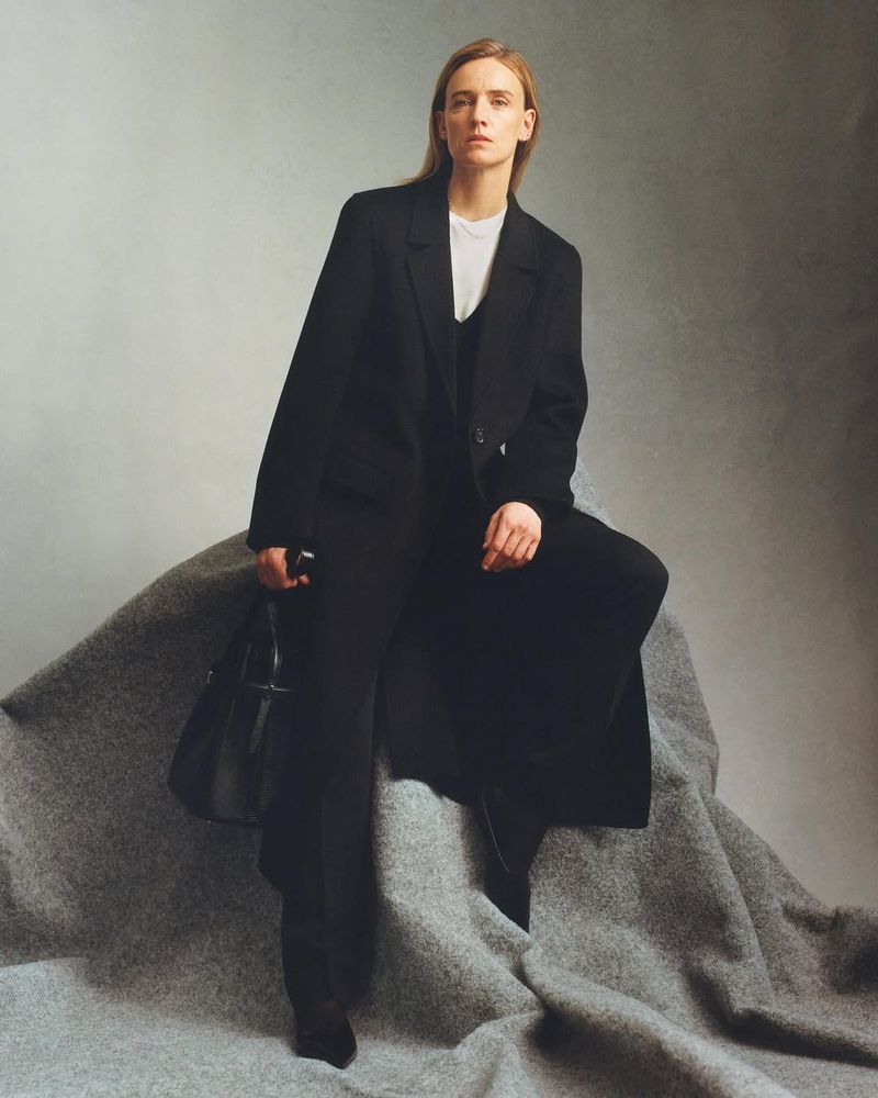 Woman in oversized black coat and pants posing on a grey fabric. She's holding a black purse, standing indoors against a neutral background.