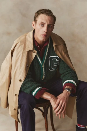 Man in a green letterman cardigan and tan trench coat sitting on a wooden chair in front of a neutral backdrop indoors.