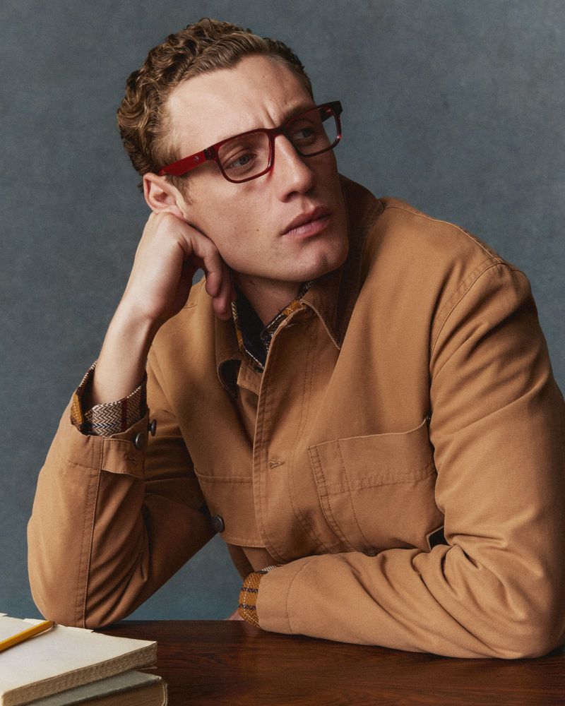 Man wearing red glasses and a tan jacket resting his hand on his chin against a textured grey background.
