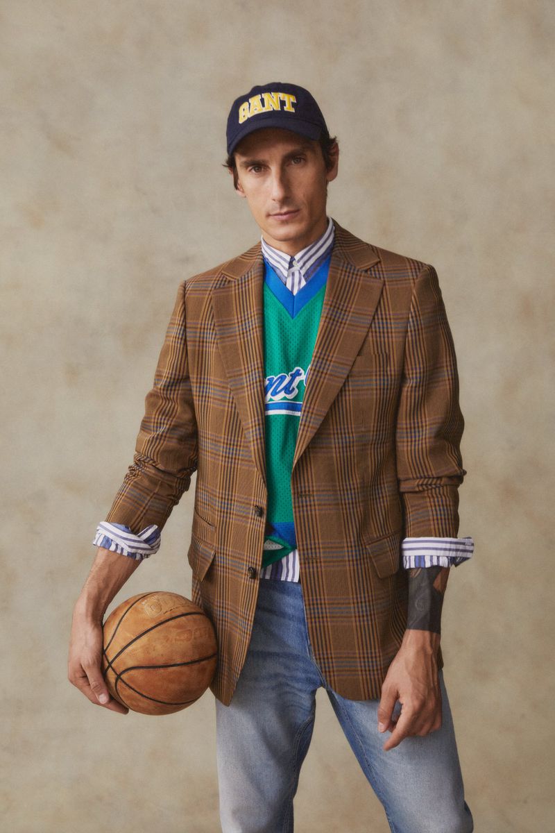 Man in blazer and cap holding basketball in studio shot, showcasing fashion and sport elements with a neutral backdrop and indoor setting.