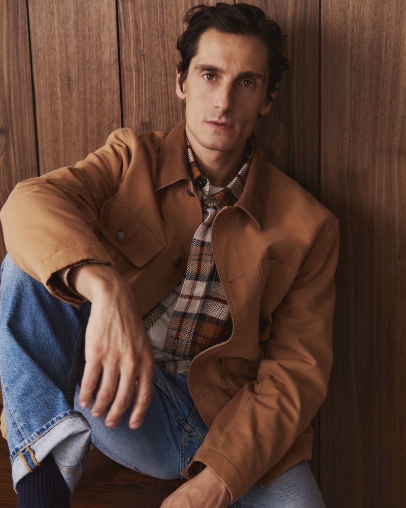 A man in a tan jacket and jeans poses against a wood background, looking directly at the camera in a casual, eye-level shot.