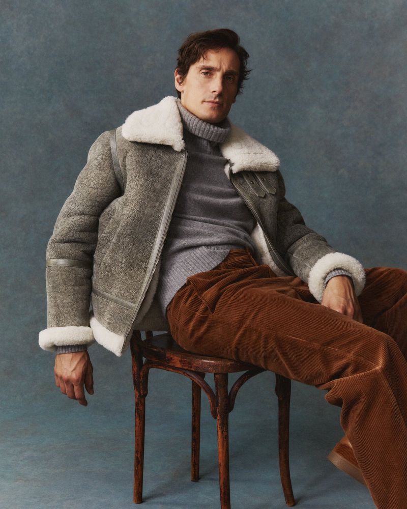 Man in grey jacket and brown corduroy pants sits on wooden chair against a blue-grey backdrop in a medium shot portrait.