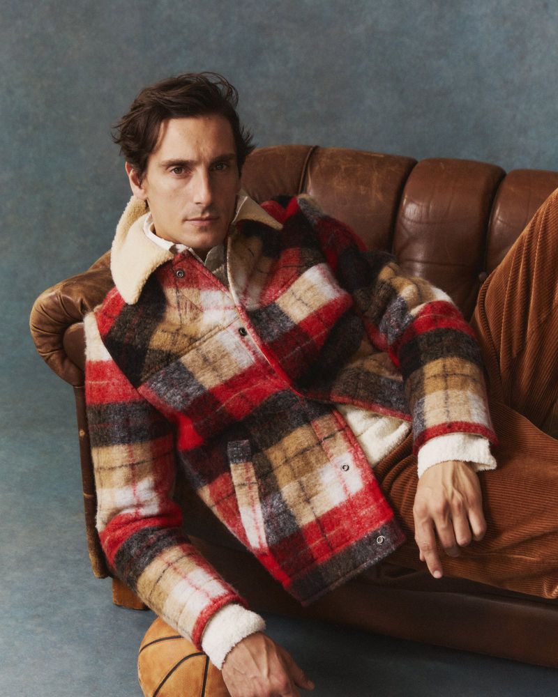 A man lounges on a leather couch wearing a plaid jacket, showcasing fashion and casual style with a basketball on the floor.