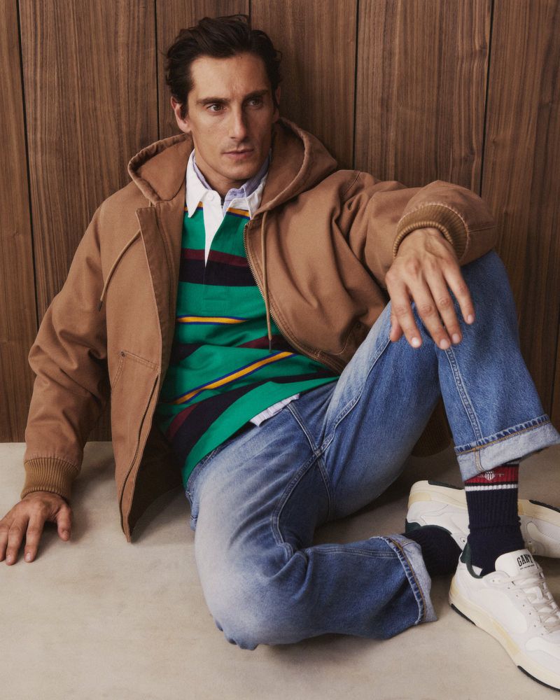 Man seated wearing a brown canvas hooded jacket, striped rugby shirt, blue jeans, and white sneakers against wood paneling