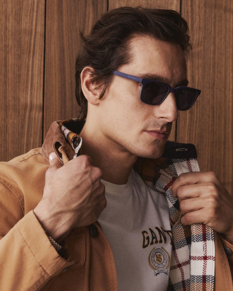 A man poses in sunglasses and a tan jacket in a medium shot against a wooden backdrop, showcasing casual fashion.