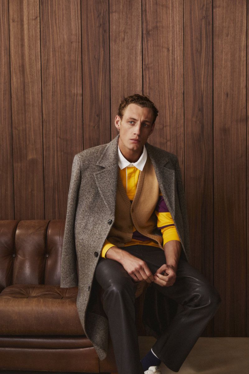 A fair-skinned adult male model sits indoors on a brown leather couch, wearing a colorful polo shirt and an overcoat.