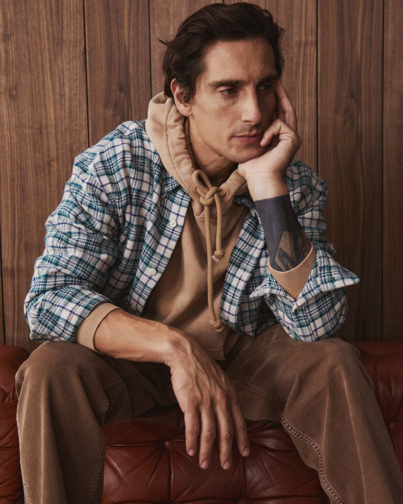 Man in hoodie and plaid shirt sitting on a leather couch. Head rests on hand. Wooden wall background. Indoor setting, medium shot.