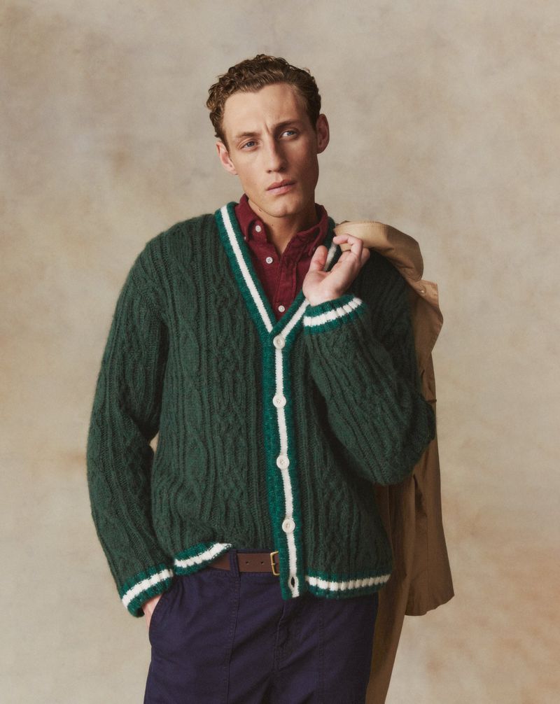 A stylish young man poses indoors wearing a green cable-knit cardigan, a red button-down shirt, and navy blue trousers with a brown belt.