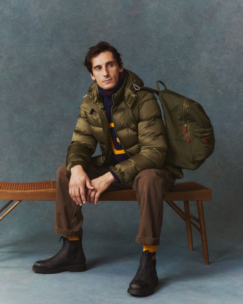 A man in a green puffer jacket and brown pants sits on a bench in a studio setting, carrying a matching backpack, fashion portrait.