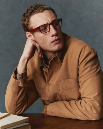 Man wearing red glasses and a tan jacket resting his hand on his chin against a textured grey background.