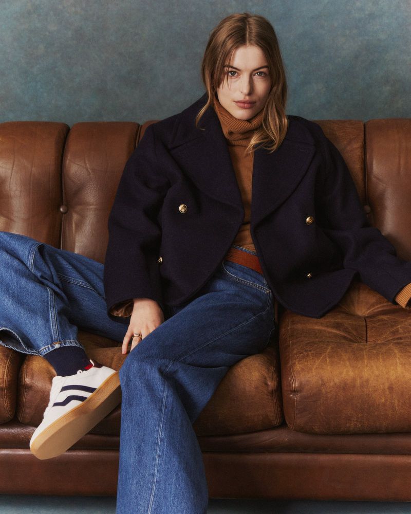 Woman seated on a brown leather sofa wearing a dark blue double-breasted jacket, blue jeans, and sneakers.