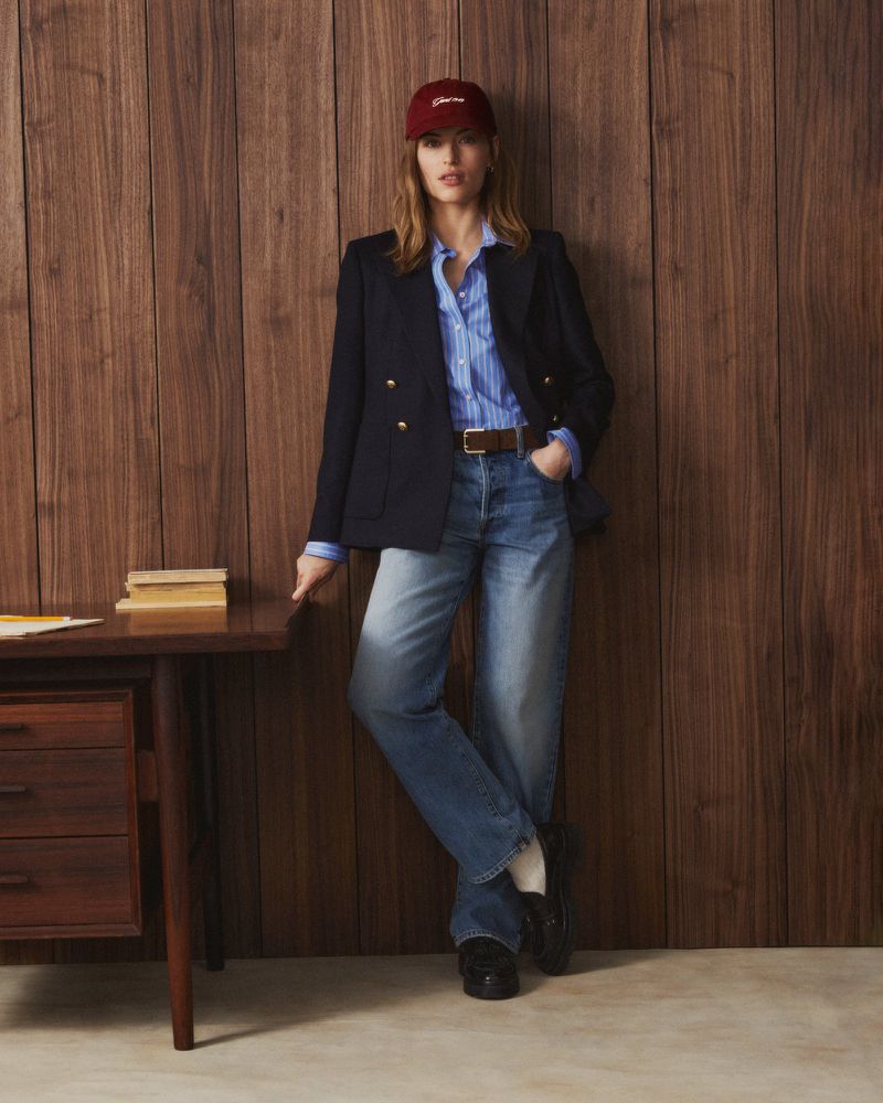 Model leaning against wood paneling wearing a navy blazer, blue striped shirt, denim jeans, and a red cap.