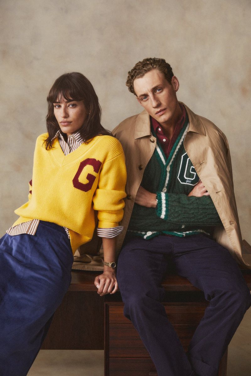 Two fashion models pose indoors, wearing sweaters with varsity letters and preppy outfits, sitting atop a vintage wood desk.