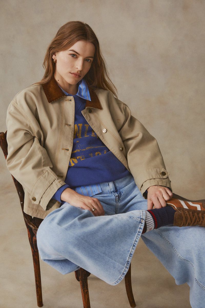 A woman with long brown hair poses indoors, wearing a tan jacket, blue top, and jeans, seated on a wooden chair with a neutral backdrop.