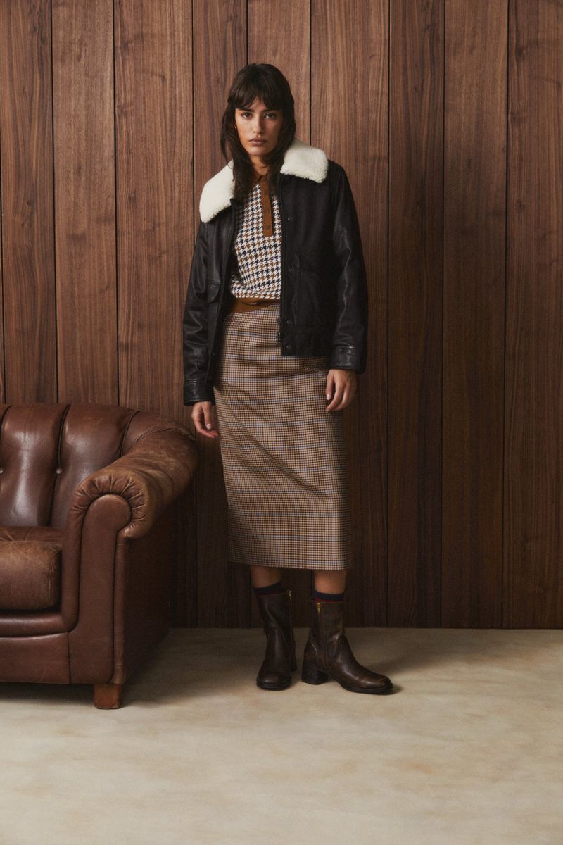 Woman in a black leather jacket with a white faux fur collar, and brown skirt, standing in front of a brown wooden wall.