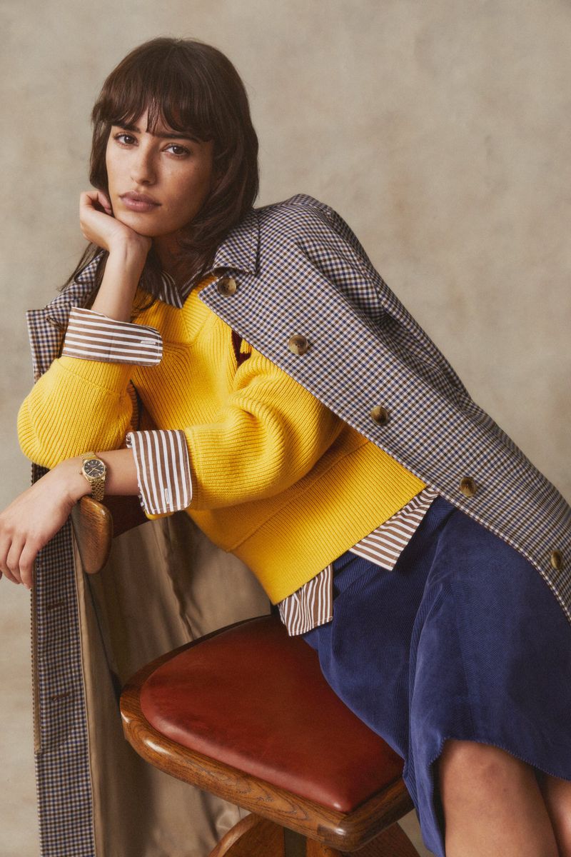A woman poses seated on a stool in a fashion studio, wearing a yellow sweater, blue skirt, and a checkered coat draped over her shoulder.