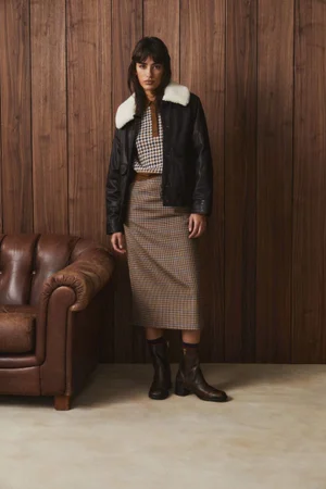 Model standing in a wood-paneled room wearing a black leather jacket with a white shearling collar and plaid skirt.