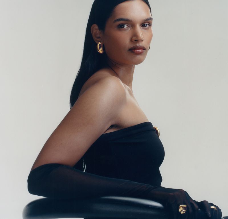 Portrait of an adult female with dark hair wearing a black top, gold earrings, gold ring, and sheer black gloves against a white backdrop.