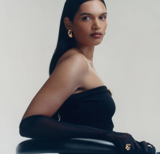 Portrait of an adult female with dark hair wearing a black top, gold earrings, gold ring, and sheer black gloves against a white backdrop.