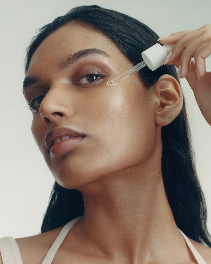 Close-up shot of a woman applying serum to her face with a dropper in a beauty or skincare context, set against a clean white background.
