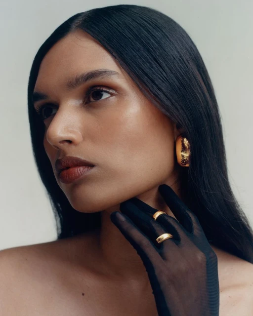 Woman wearing gold hoop earrings and sheer black gloves poses in a studio with a neutral background in a beauty fashion style.
