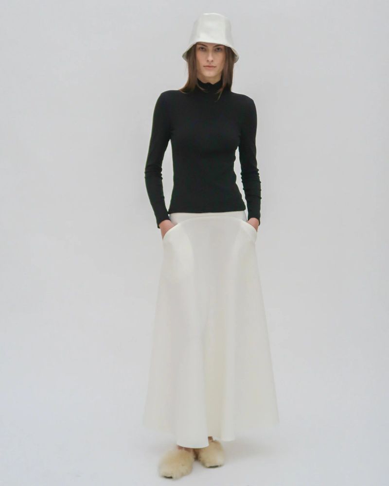 Full-length shot of a woman in a black long-sleeve shirt and white skirt with a matching bucket hat, standing in a white studio.