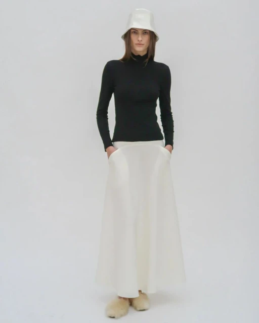 Full-length shot of a woman in a black long-sleeve shirt and white skirt with a matching bucket hat, standing in a white studio.
