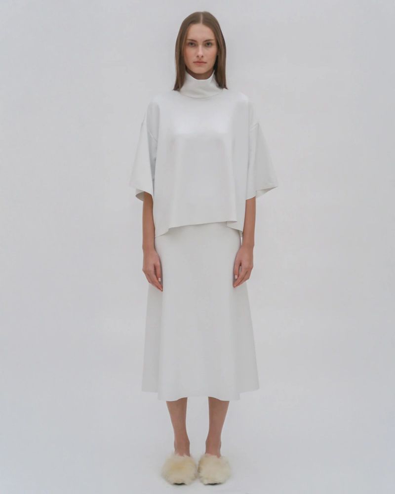 Studio shot of a model in a white turtleneck top and midi skirt, posing against a white background. She is wearing furry slippers.